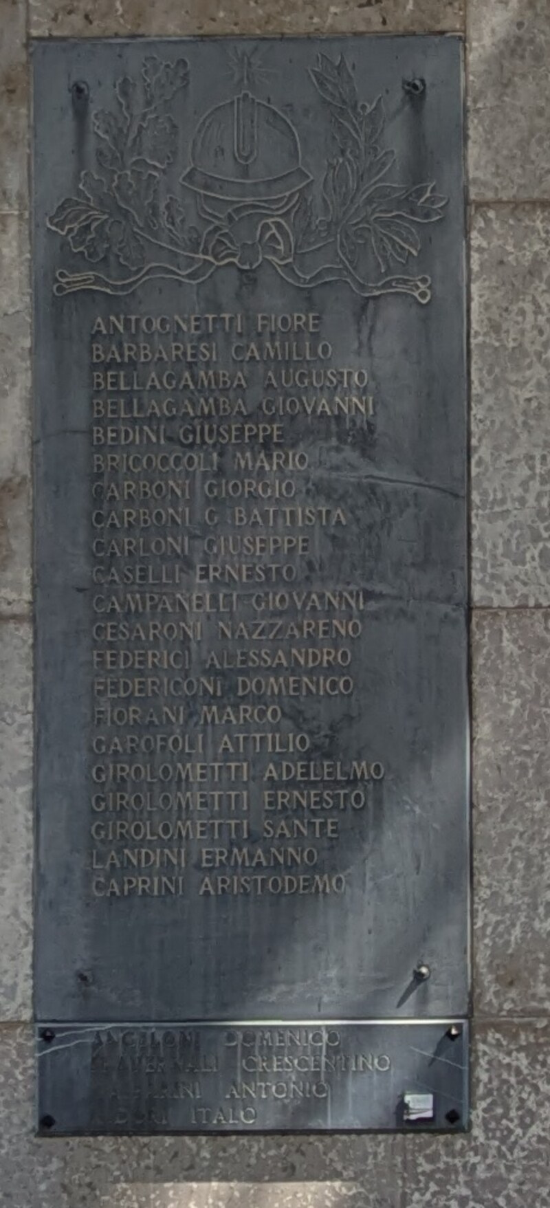 Lapide scura verticale con in alto un elmetto in rilievo fra rami di quercia e alloro annodati da un nastro; sotto, ventuno righe di nomi in lettere maiuscole chiare. In basso, staccata, una striscia più piccola con quattro righe quasi cancellate.