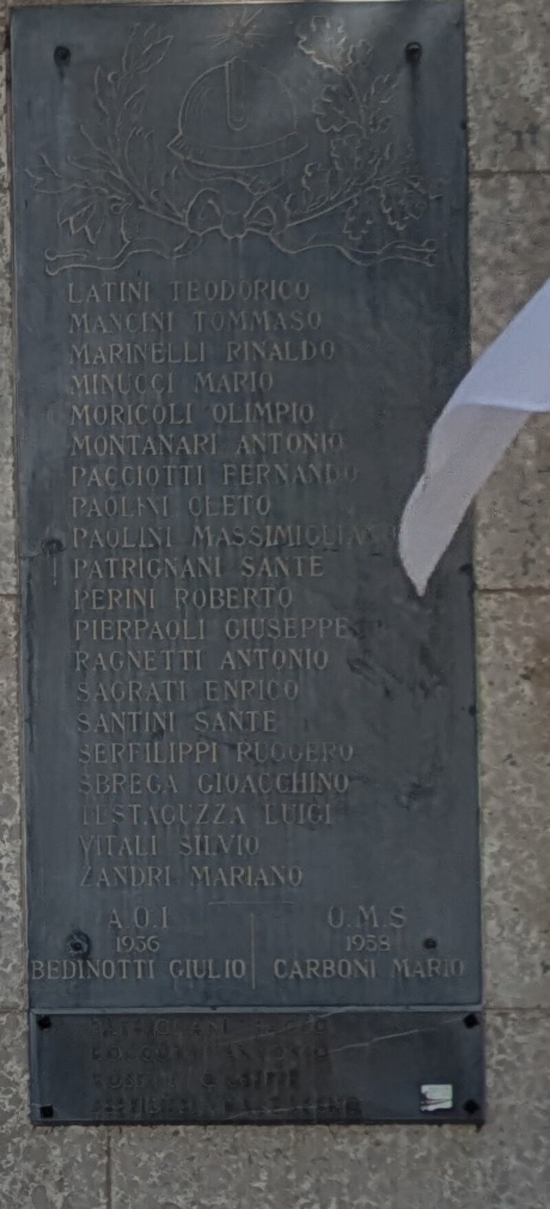 Seconda lapide scura verticale, uguale alla prima, con l'elmetto in rilievo in alto e venti righe di nomi; in fondo, separate da una linea, due sigle con una data e un nome ciascuna. Una bandiera bianca e azzurra ne copre in parte l'angolo destro.