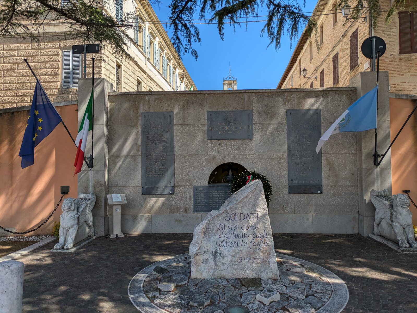 Un muro basso in pietra chiara, con sei lapidi scure di bronzo o metallo. Al centro in alto una lapide con uno stemma e tre righe di iscrizione; ai due lati due lapidi verticali più grandi, coperte di nomi in lettere chiare, e sotto ciascuna delle due, staccata e avvitata a parte, una striscia orizzontale piccola con quattro righe quasi cancellate; sotto quella centrale, una lapide orizzontale con due colonne di nomi, il cui angolo in basso a destra è nascosto. Al centro del muro una nicchia ad arco, davanti alla quale pende una corona d'alloro. Ai lati del muro due pilastri con bandiere — europea e italiana a sinistra, una bandiera azzurra e bianca a destra — e due leoni alati di pietra alla base; da ciascun pilastro una catena parte verso l'esterno, lungo il perimetro, e non verso l'altro leone. Davanti alla lapide di sinistra, appoggiato al muro, un leggìo di pietra chiara con sopra un pannello stampato. Davanti al muro, al centro, una pietra grezza chiara con un'iscrizione azzurra. Sopra il muro si vedono un campanile a vela con la croce, i tetti delle case e una bandiera verde e bianca su un palazzo dietro.