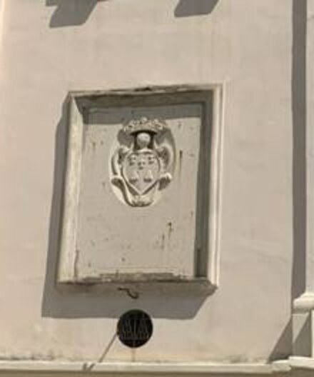 Dettaglio della lapide. Dentro una cornice di pietra chiara, incassata nel muro, uno stemma scolpito in rilievo occupa la parte alta: uno scudo sagomato con volute laterali, sormontato da una corona. Sotto lo stemma, per circa due terzi dell'altezza del riquadro, la superficie appare liscia e priva di incisioni. In basso, fuori dalla cornice, un oculo tondo con grata.