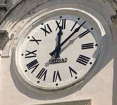 Dettaglio del quadrante dell'orologio: disco bianco con le ore in numeri romani neri e un punto fra l'una e l'altra, due lancette nere che segnano quasi mezzogiorno. Sotto il perno delle lancette, in stampatello nero, la scritta TREBINO.